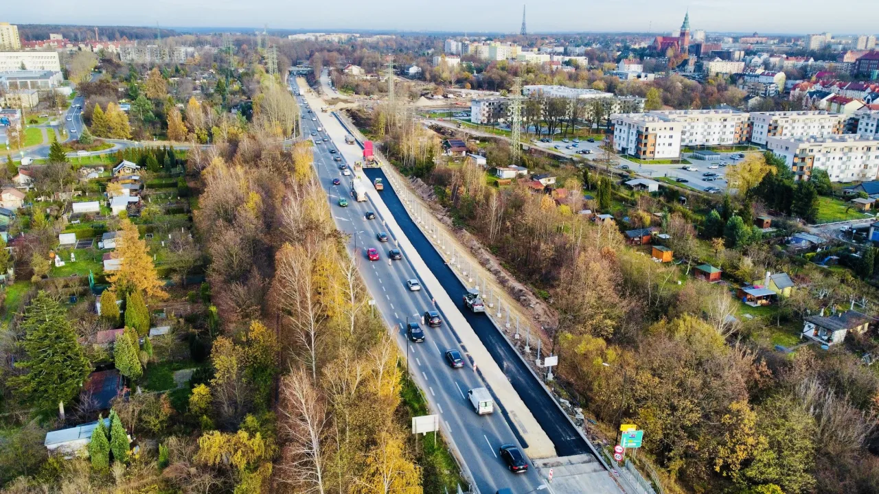Budowa drogi krajowej 88 w otoczeniu jesiennej zieleni i zabudowań. Ruch samochodowy odbywa się na częściowo ukończonej trasie.