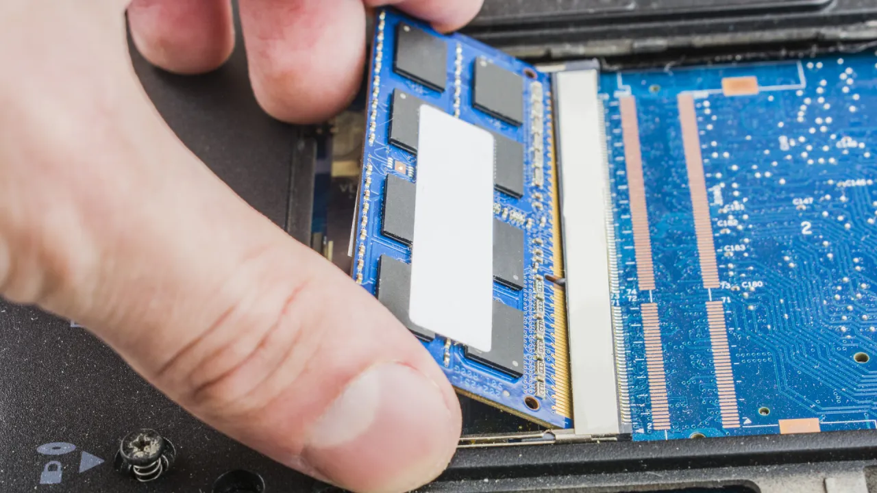 Dłoń montuje moduł RAM, przygotowując upgrade laptopa. Widać niebieską płytkę drukowaną z czarnymi kośćmi pamięci.
