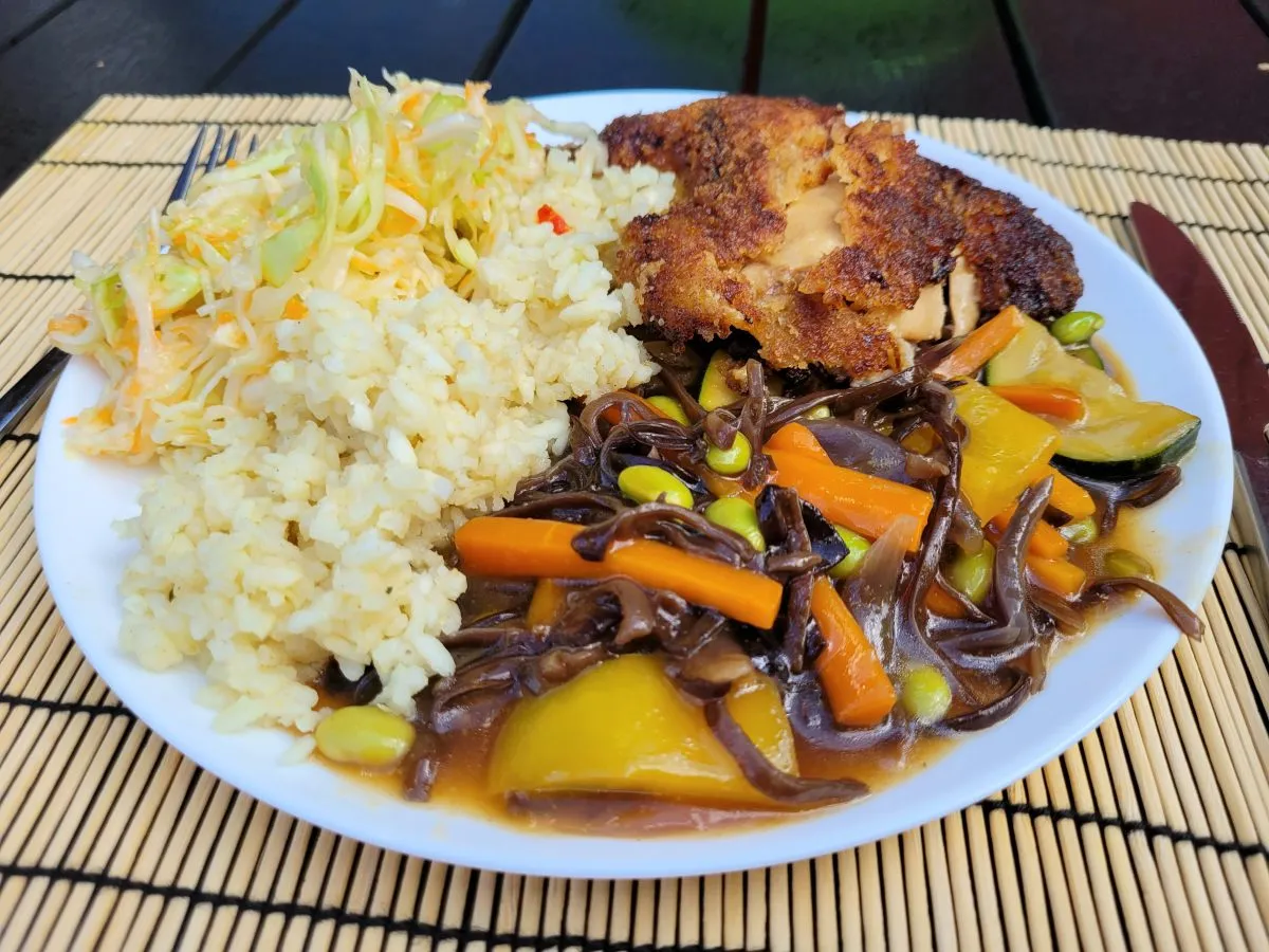 Talerz z ryżem, sur&oacute;wką i panierowanym kotletem, obok kolorowe warzywa w sosie sojowym: marchewka, cukinia, papryka i edamame.