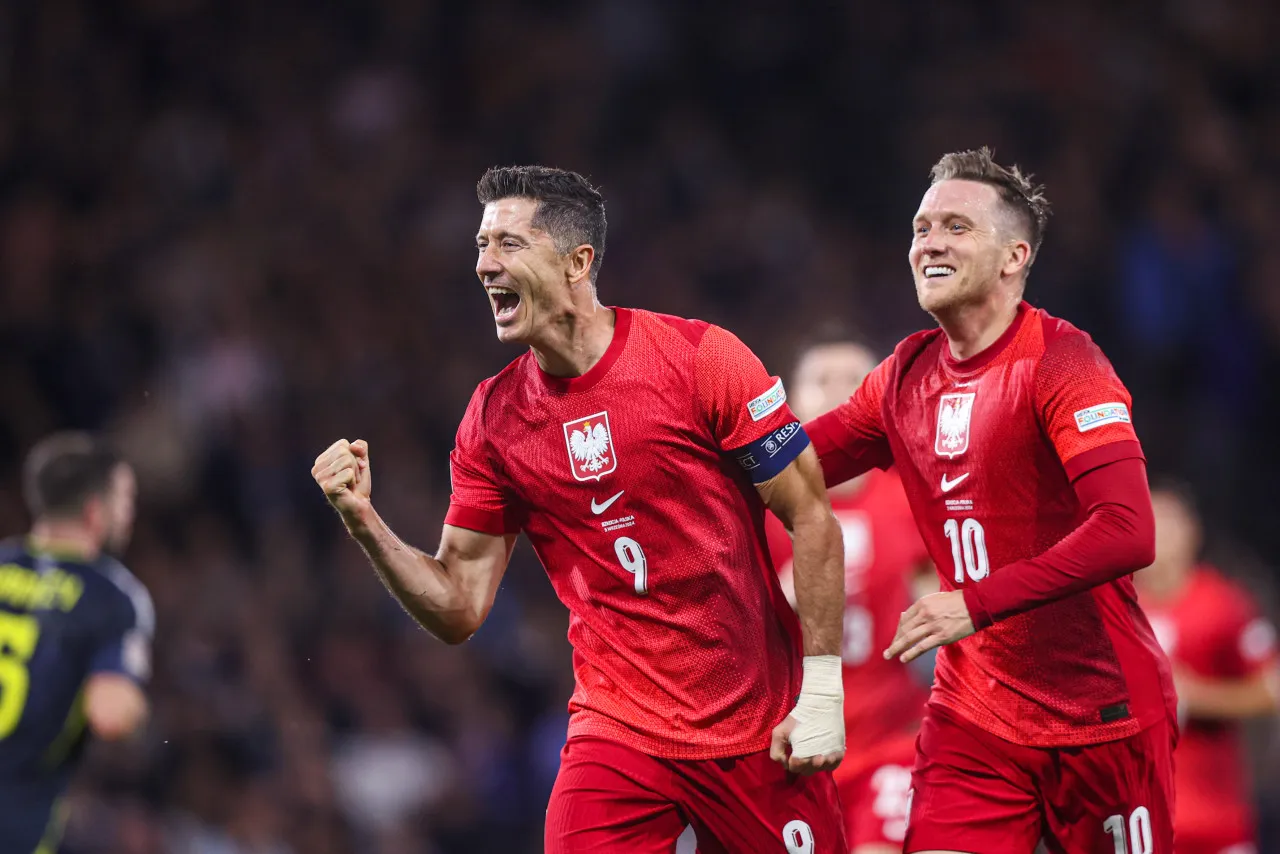 Dw&oacute;ch piłkarzy reprezentacji Polski w czerwonych strojach świętuje bramkę. Widać ich radość, gdy grają w piłkę nożną.