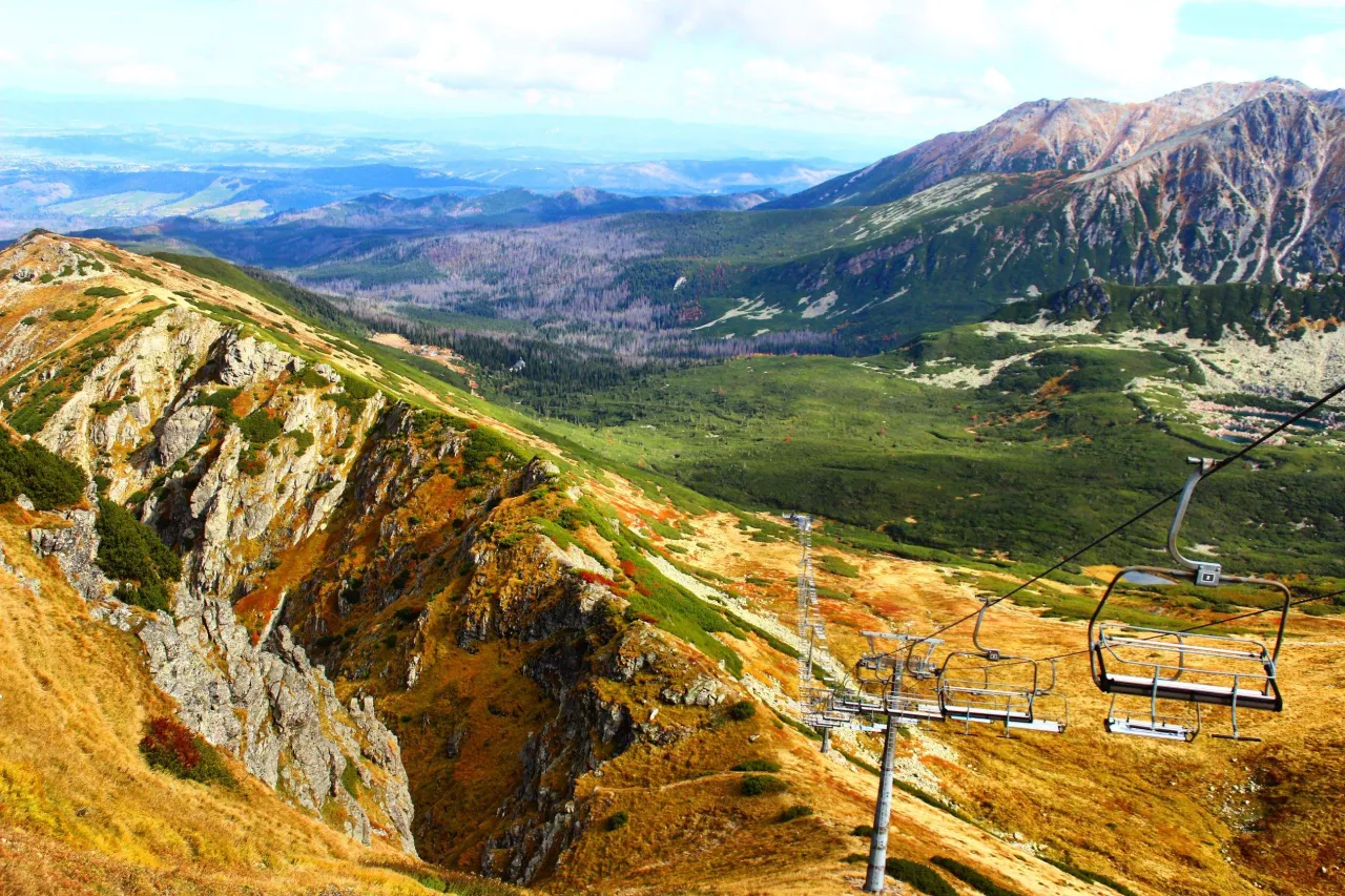 Jesień na szlaku na Kasprowy Wierch. Kolejka krzesełkowa wjeżdża na zbocze pokryte złotą trawą i zieloną kosodrzewiną. W oddali widać pasma g&oacute;rskie.