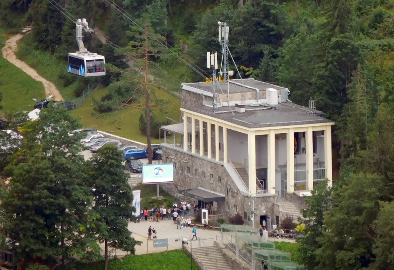 Kolejka na Kasprowy Wierch wjeżdża na g&oacute;rę. Obok budynku stacji widoczny jest parking i ludzie czekający na wejście. Sprawdź wyciąg na Kasprowy Wierch cennik.