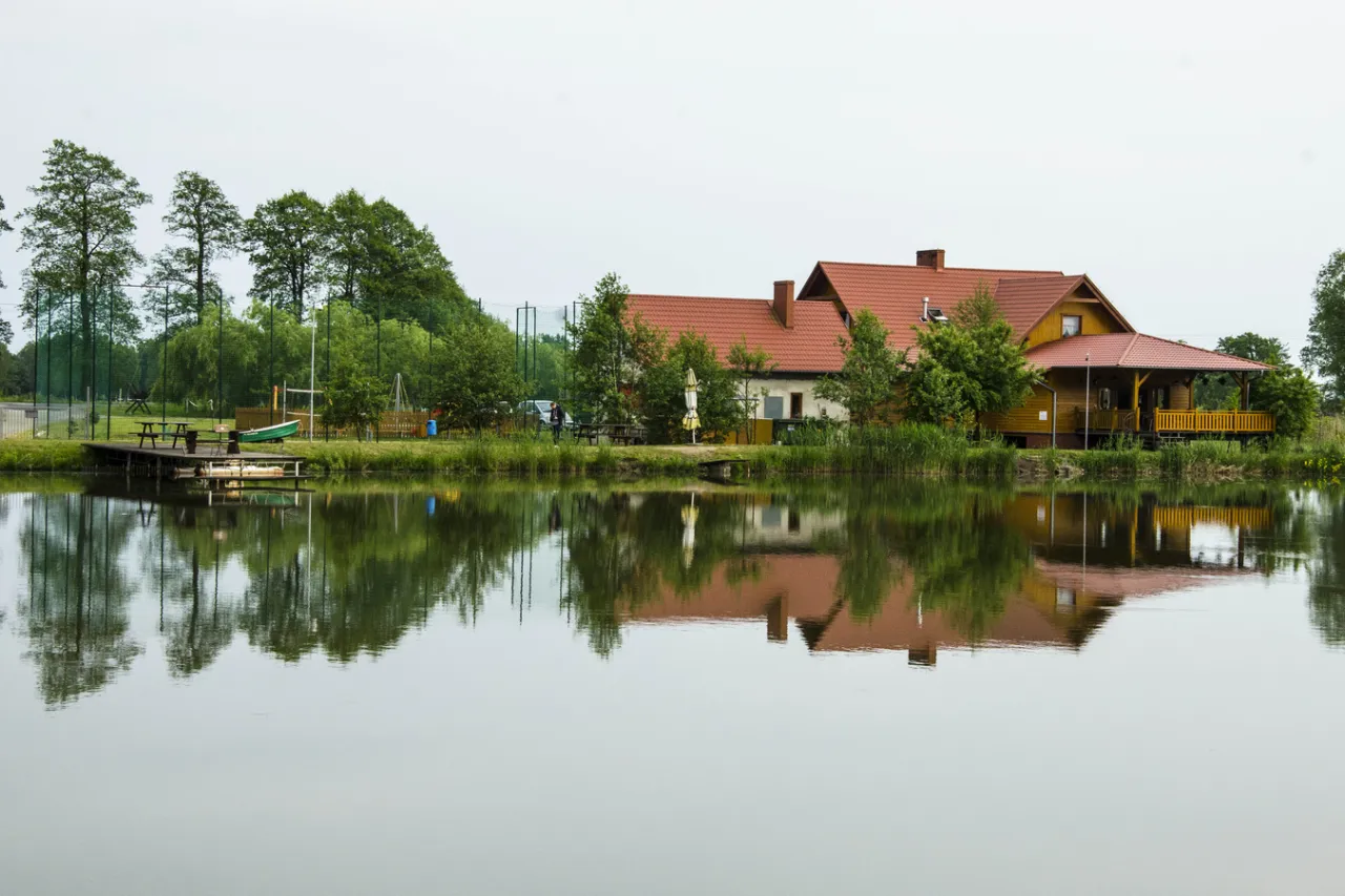 U Bartka smażalnia ryb i łowisko. Drewniany budynek z czerwoną dachem, pomost, łódka i odbicie w spokojnej wodzie.