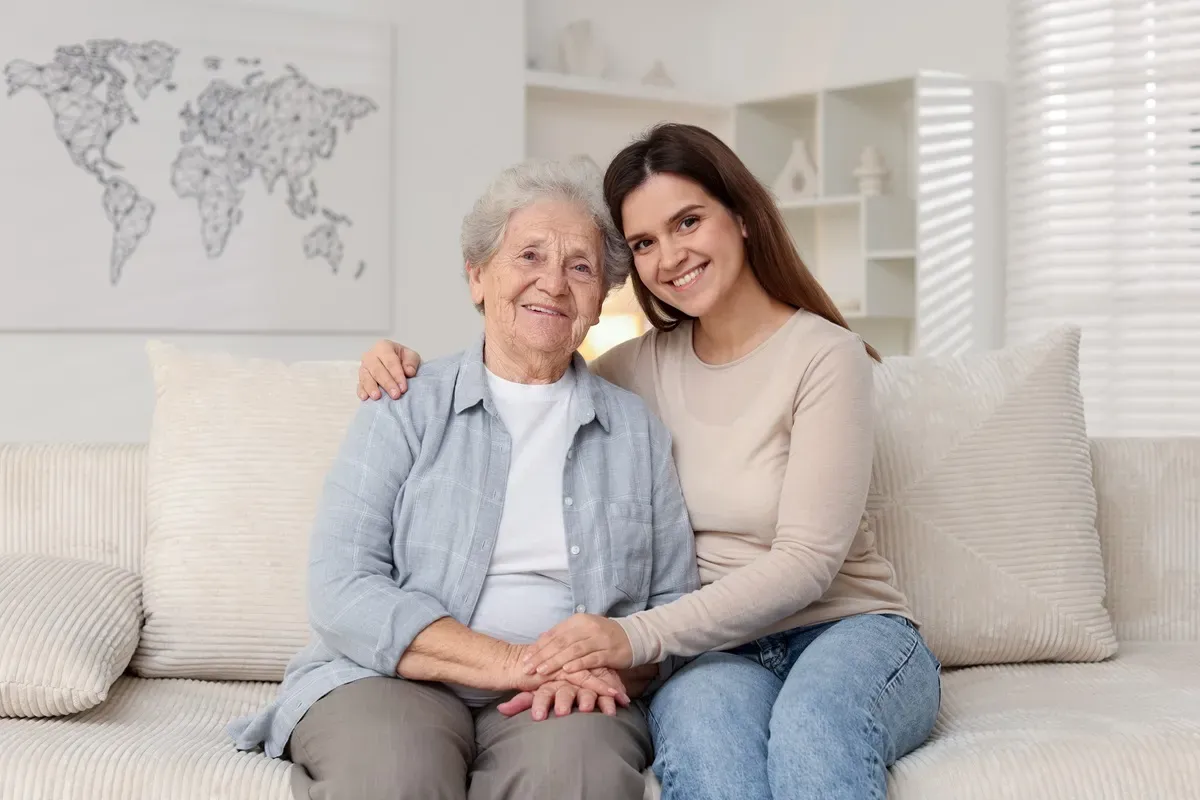 Uśmiechnięta młoda kobieta obejmuje starszą panią. Obraz symbolizuje wsparcie i troskę, kluczowe przy ubieganiu się o zasiłek za opiekę nad osobą starszą.