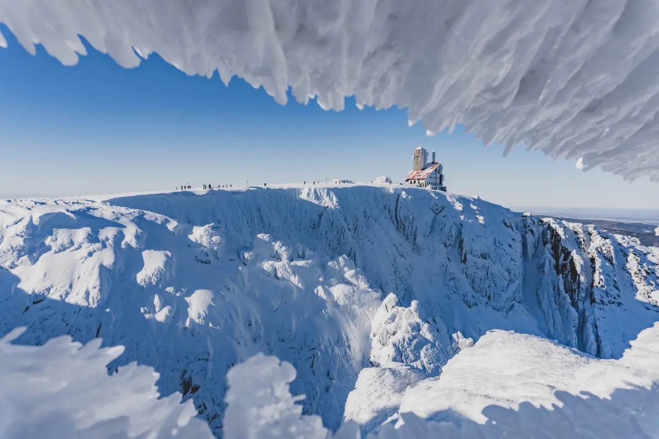 Zimowy krajobraz z lodowymi sopelkami w kadrze. Na szczycie góry, pokrytej śniegiem, stoi schronisko pod Łabskim Szczytem. W oddali widać turystów.