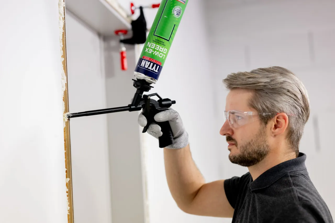 Mężczyzna w okularach ochronnych i rękawicach piankuje futrynę, pokazując, jak zamontować drzwi wewnętrzne.