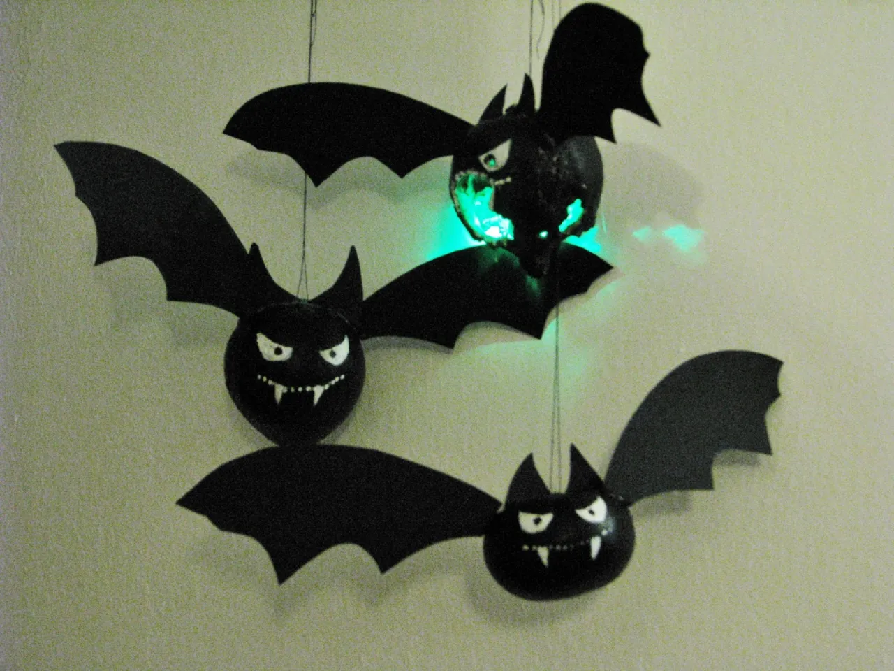 Jak zrobić ozdoby na Halloween? Trzy czarne nietoperze z wyciętymi skrzydłami i strasznymi minami wiszą na ścianie. Jeden świeci na zielono.