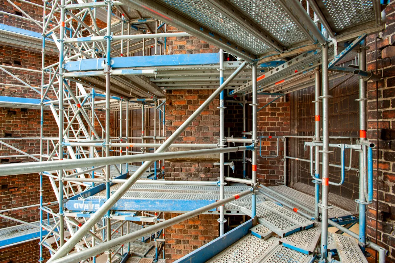 Skomplikowana konstrukcja rusztowania aluminiowego przy ceglanej ścianie. Widać elementy do montażu, jak złożyć rusztowanie aluminiowe.