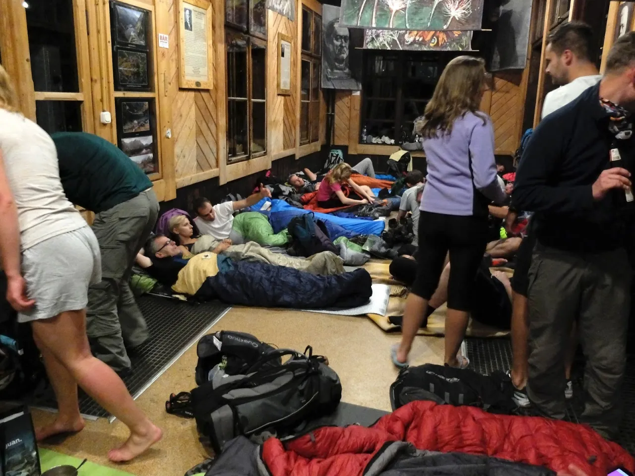 Ludzie śpią na podłodze w schronisku turystycznym, otoczeni śpiworami i plecakami.