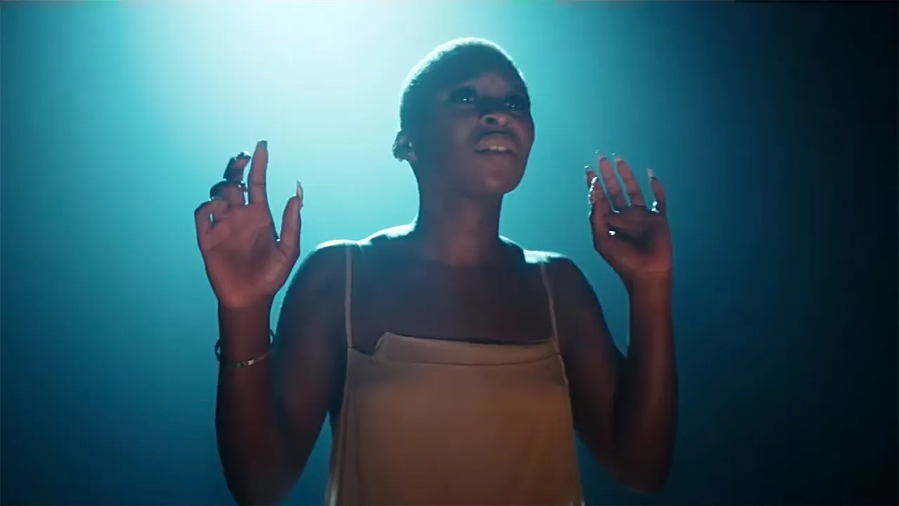 Cynthia Erivo stand up, wznosi ręce w g&oacute;rę, w świetle reflektor&oacute;w.