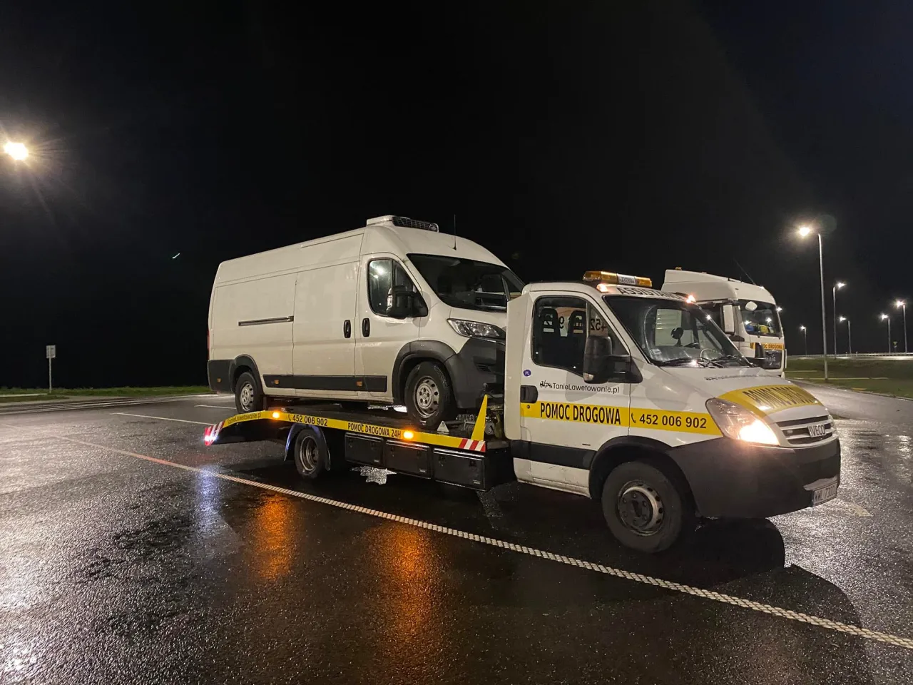 Pomoc drogowa holuje białą furgonetkę na drodze ekspresowej. Czy można holować samochód na drodze ekspresowej? Tak, gdy potrzebna jest pomoc.