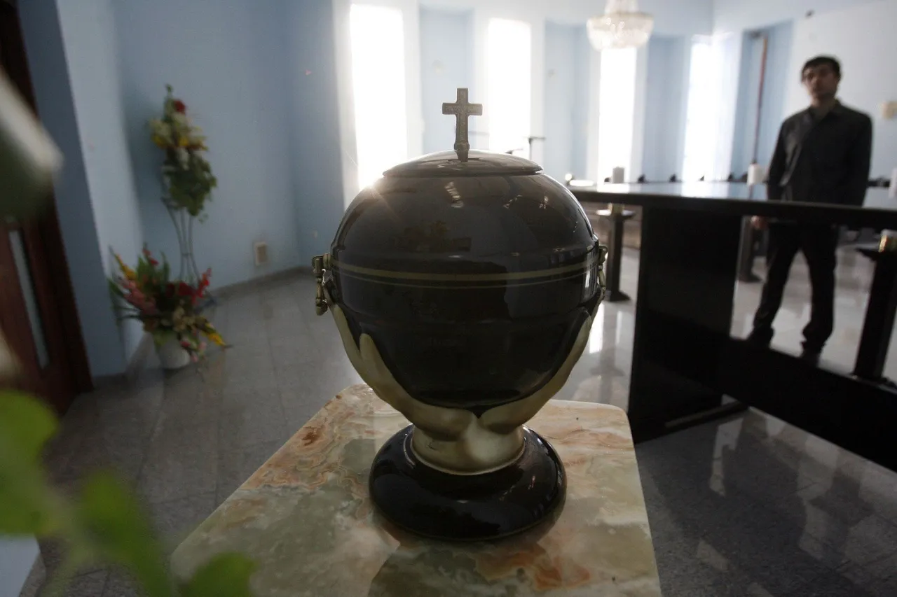 Ciemna urna z krzyżem na szczycie, stojąca na marmurowym postumencie. Czy kremacja jest grzechem? W tle mężczyzna i kwiaty.