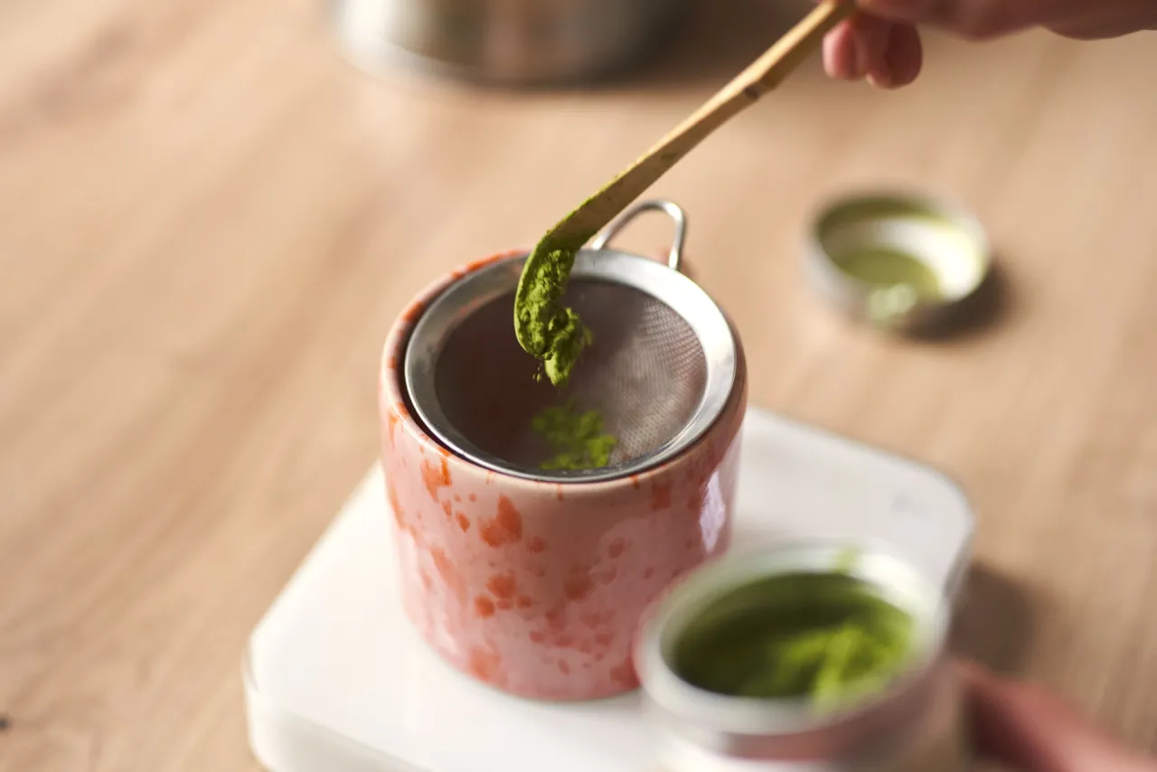 Przygotowanie matcha jaką najlepsza: przesiewanie zielonego proszku do różowego naczynia.