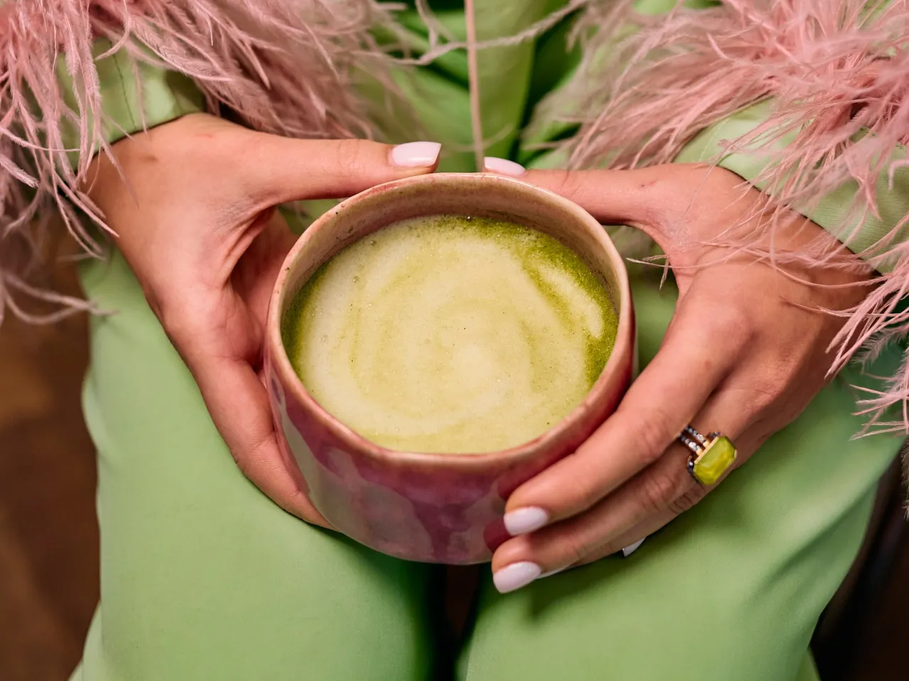 Dłonie trzymają kubek z zieloną matchą, zastanawiając się, czy matcha odchudza. R&oacute;żowe pi&oacute;ra dodają elegancji.