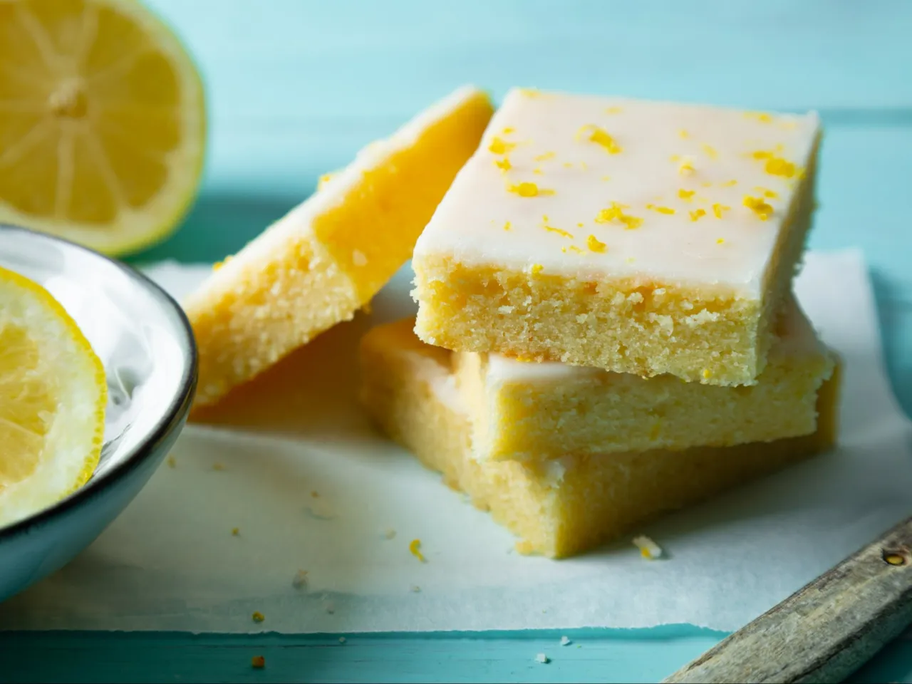 Ciasto mocno cytrynowe przepis: puszyste kwadraty z lukrem i skórką cytrynową, obok plasterki cytryny.
