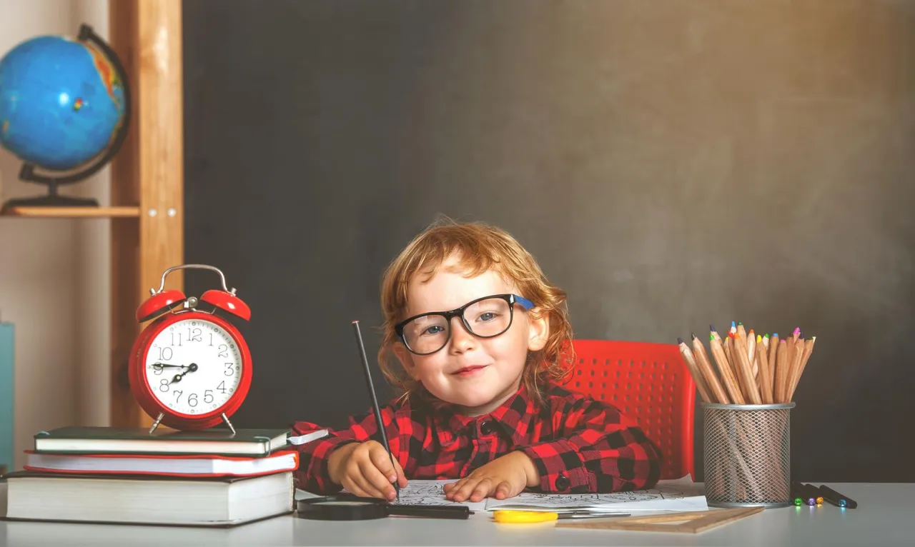 Mały uczeń z okularami rysuje przy biurku. Obok budzik i kredki. Czy wniosek o dofinansowanie do żłobka trzeba składać co miesiąc?
