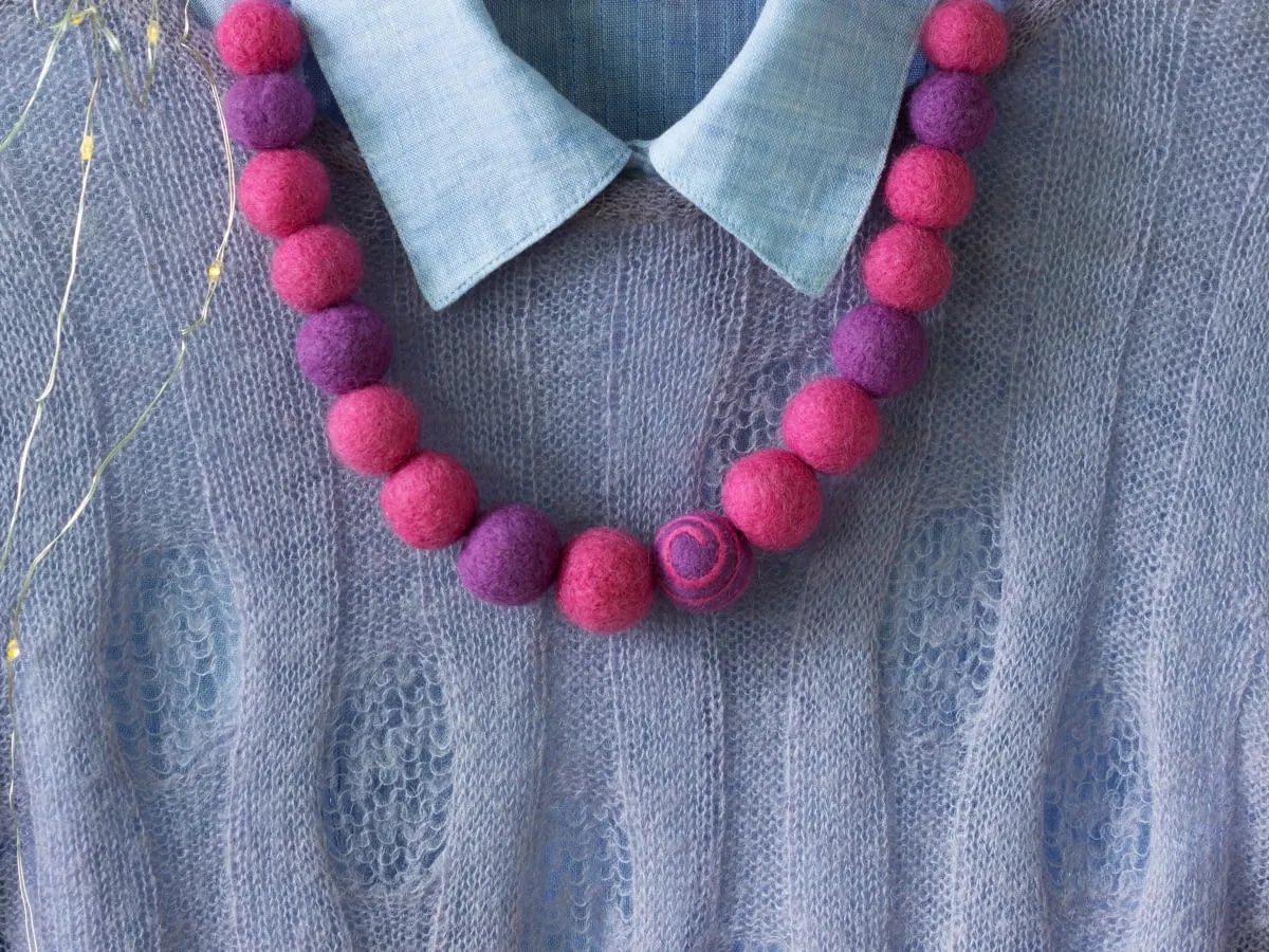 Naszyjnik z kulek z wełny czesankowej w kolorach fuksji i fioletu, ozdabia błękitną, ażurową tkaninę.