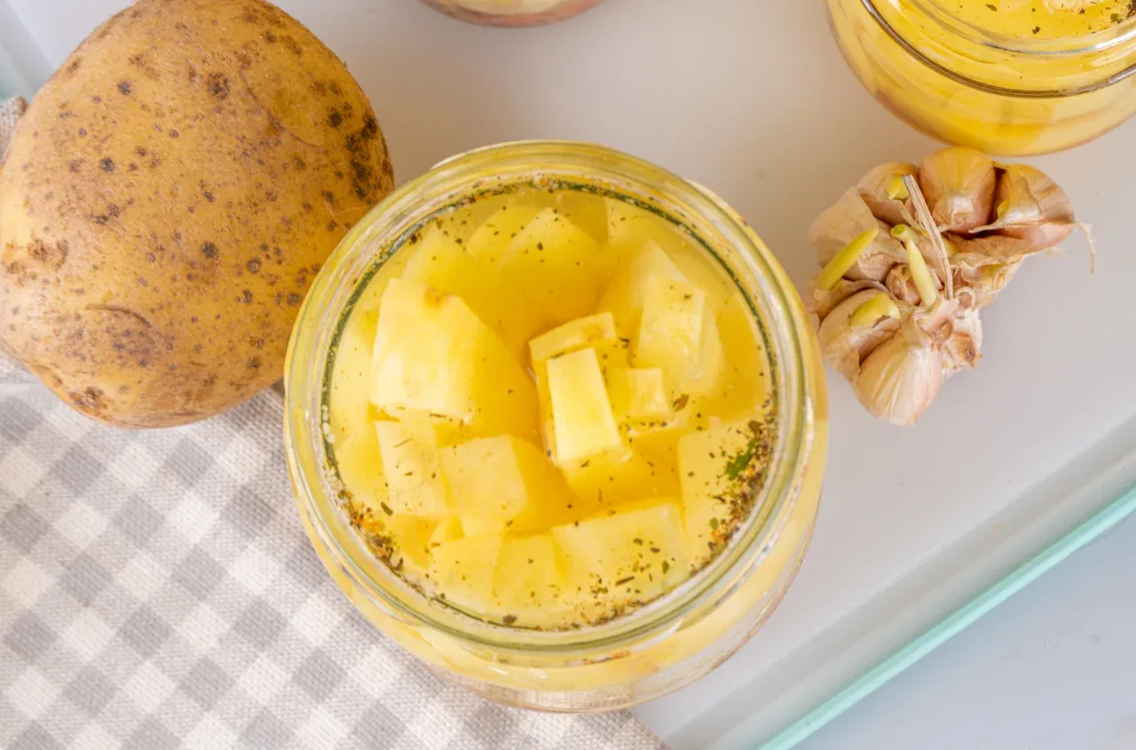 Słoik z pokrojonymi ziemniakami w zalewie, obok surowy ziemniak i czosnek. Czy obrane ziemniaki mogą stać w wodzie? Tak, w tej formie.
