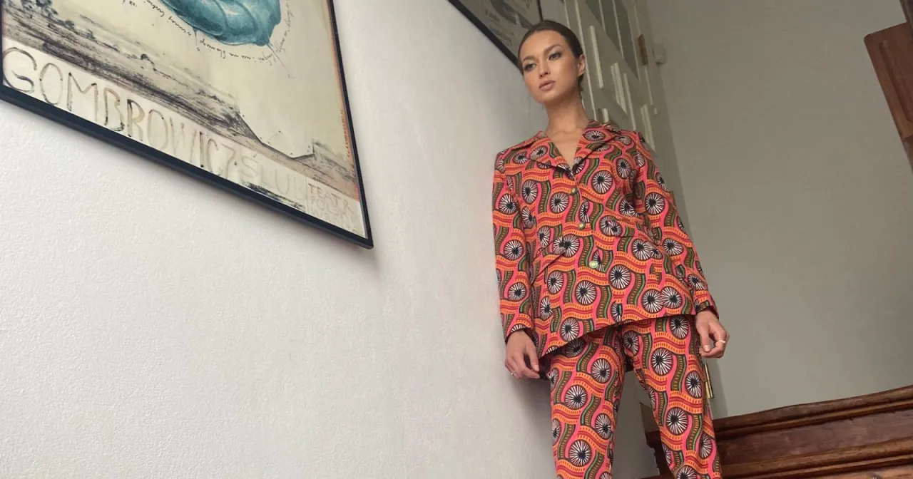 Kobieta w kolorowym, wzorzystym garniturze stoi na schodach. Obok wisi plakat z napisem 