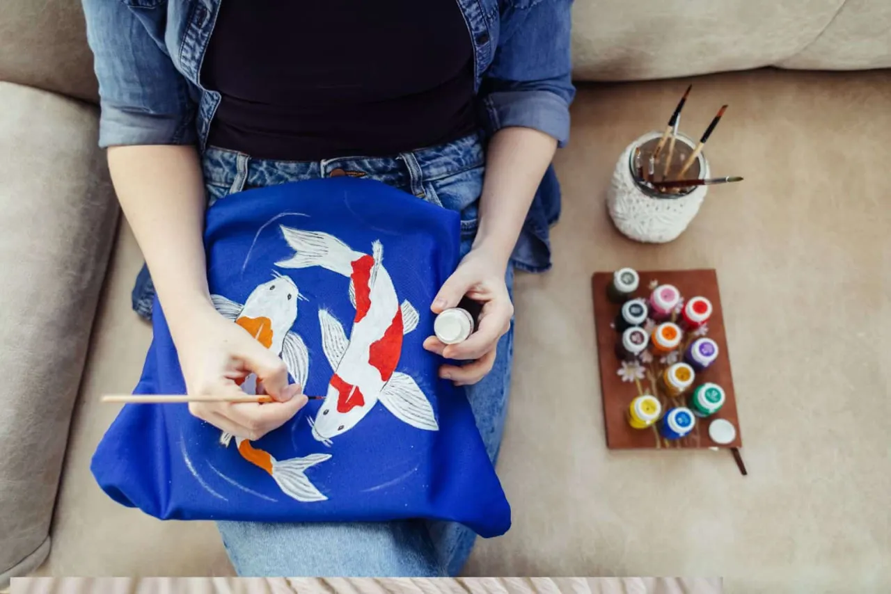 Osoba tworzy malowanie na tkaninie, dodając detale do karpi koi na niebieskiej torbie.
