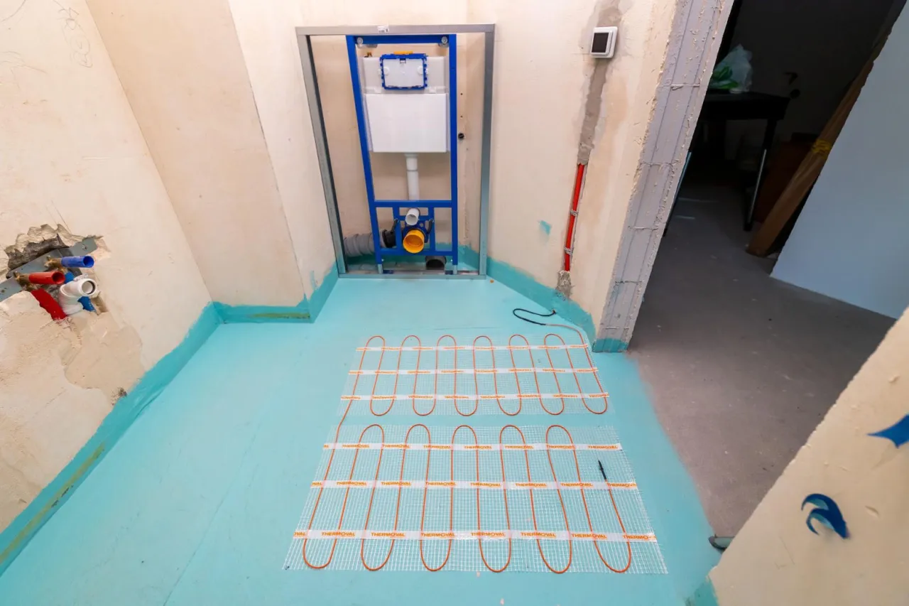 Instalacja ogrzewania podłogowego w łazience. Widać stelaż podtynkowy do WC, przewody wodne i matę grzewczą. To część prac budowlanych i remontowych.