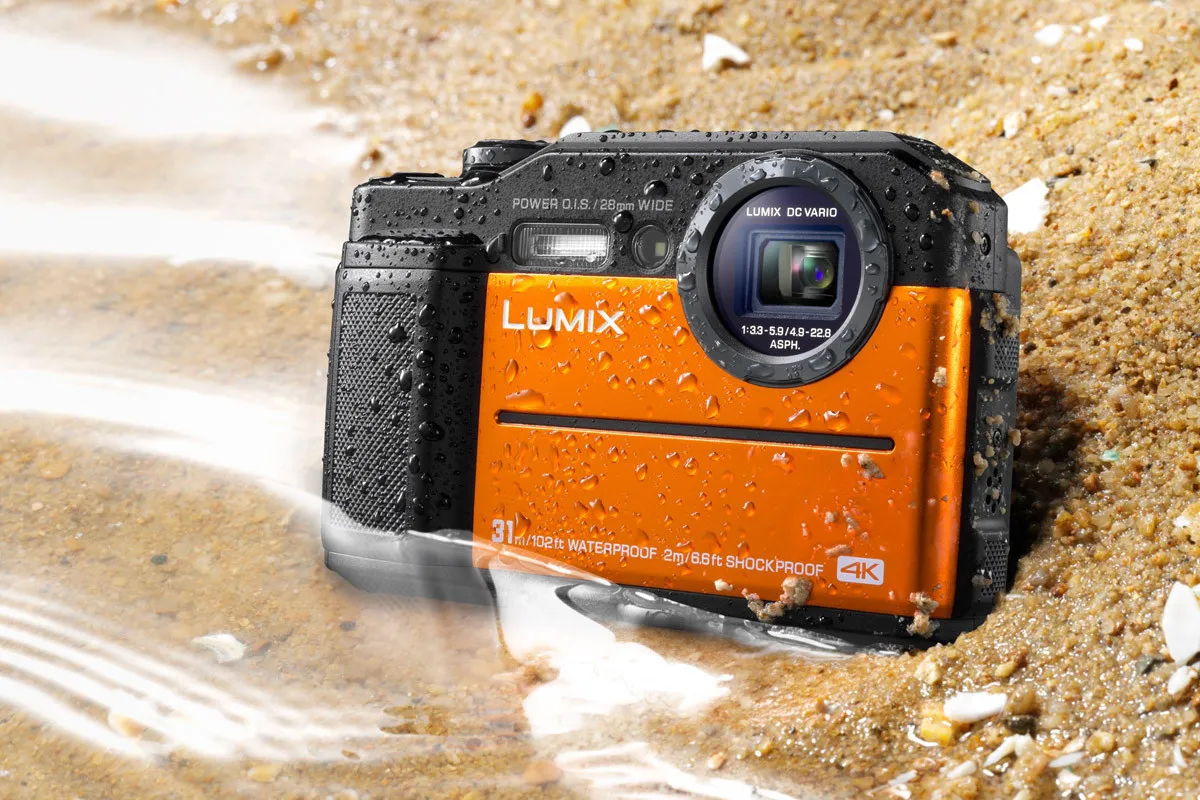 Pomarańczowy aparat Lumix DC Vario, idealny jako aparat do zdjęć podwodnych, leży na mokrym piasku. Jest wodoodporny do 31m i wstrząsoodporny.