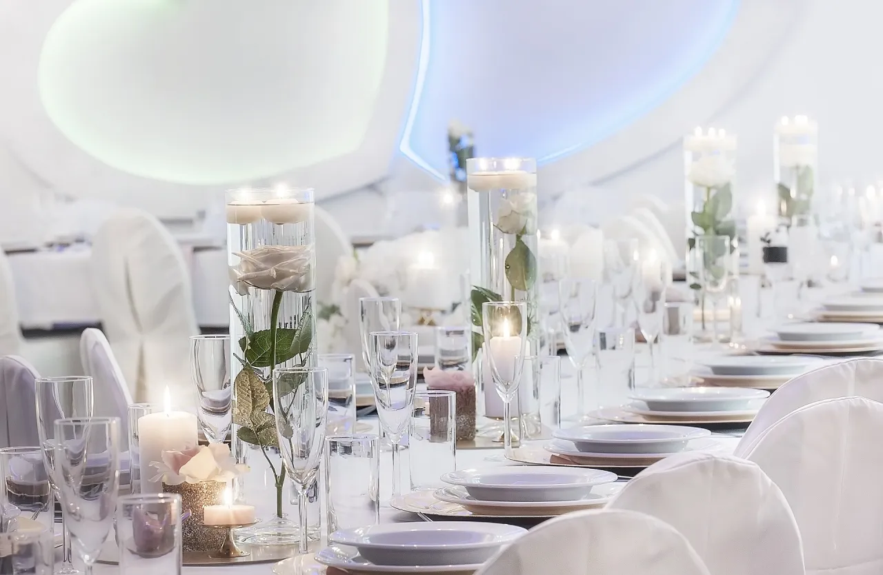 Elegancka dekoracja sali weselnej inspiracje: stoły nakryte białymi obrusami, szkło, świece i róże w wysokich wazonach.
