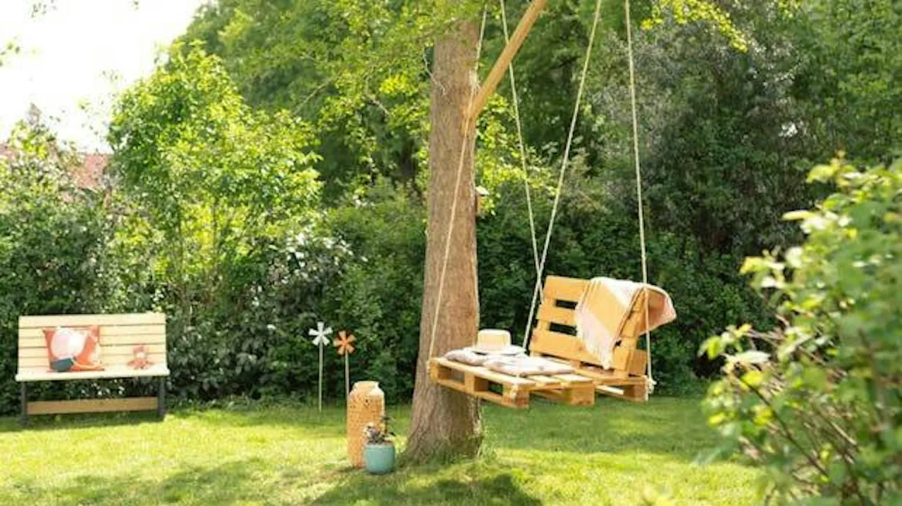 Huśtawka z drewna z palet, zawieszona na drzewie, z poduszkami i kocem. Obok ławka i ozdoby ogrodowe. Idealna inspiracja, jak zrobić huśtawkę z drewna.