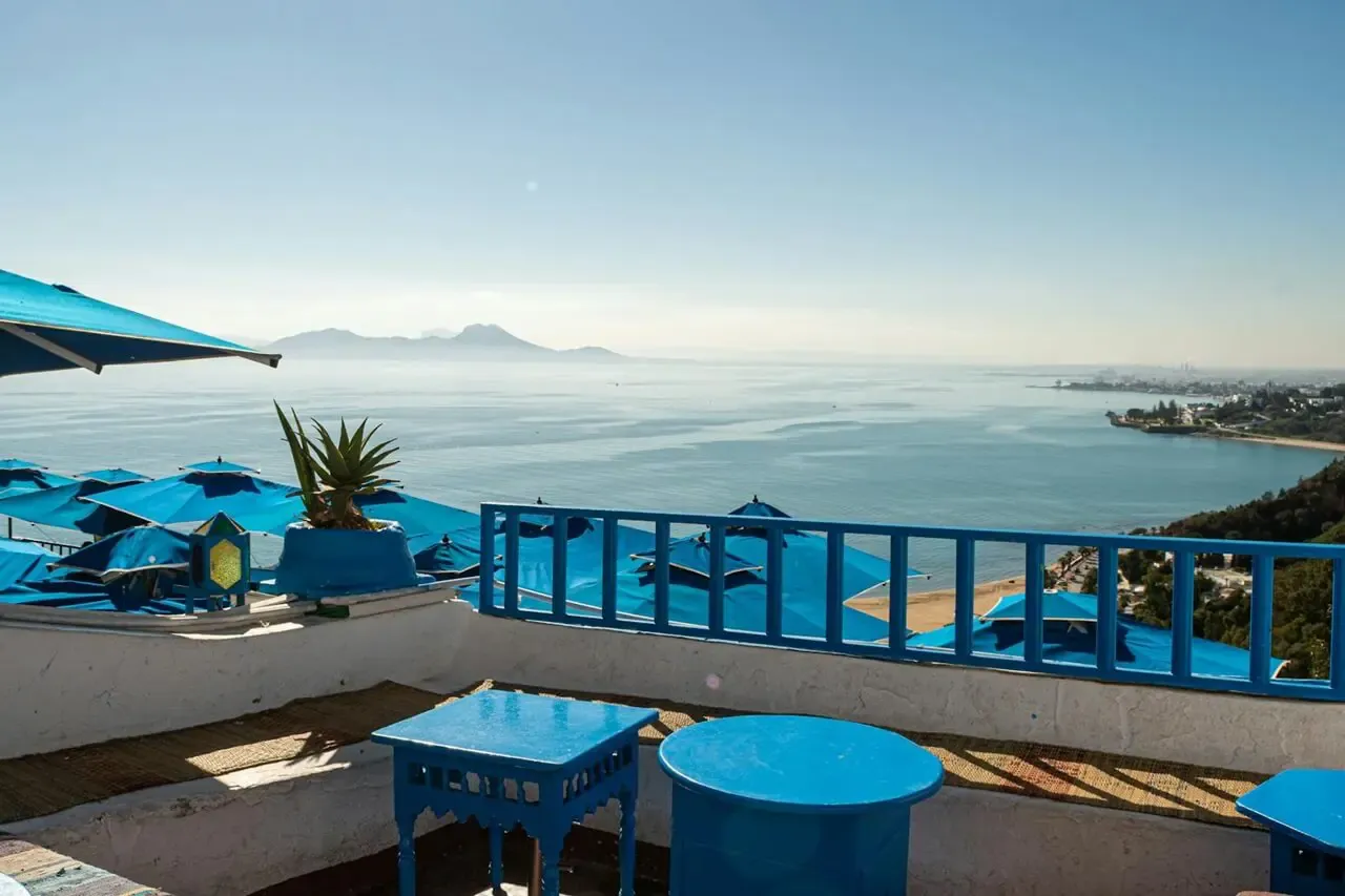 Widok na morze z niebieskimi parasolami i stolikami. Marzysz o podr&oacute;ży? Sprawdź, ile trwa lot do Tunezji z Katowic!