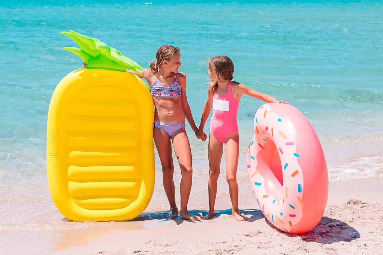 Dwie dziewczynki stoją na plaży obok dmuchanego materaca w kształcie ananasa i r&oacute;żowej dmuchanej opony.