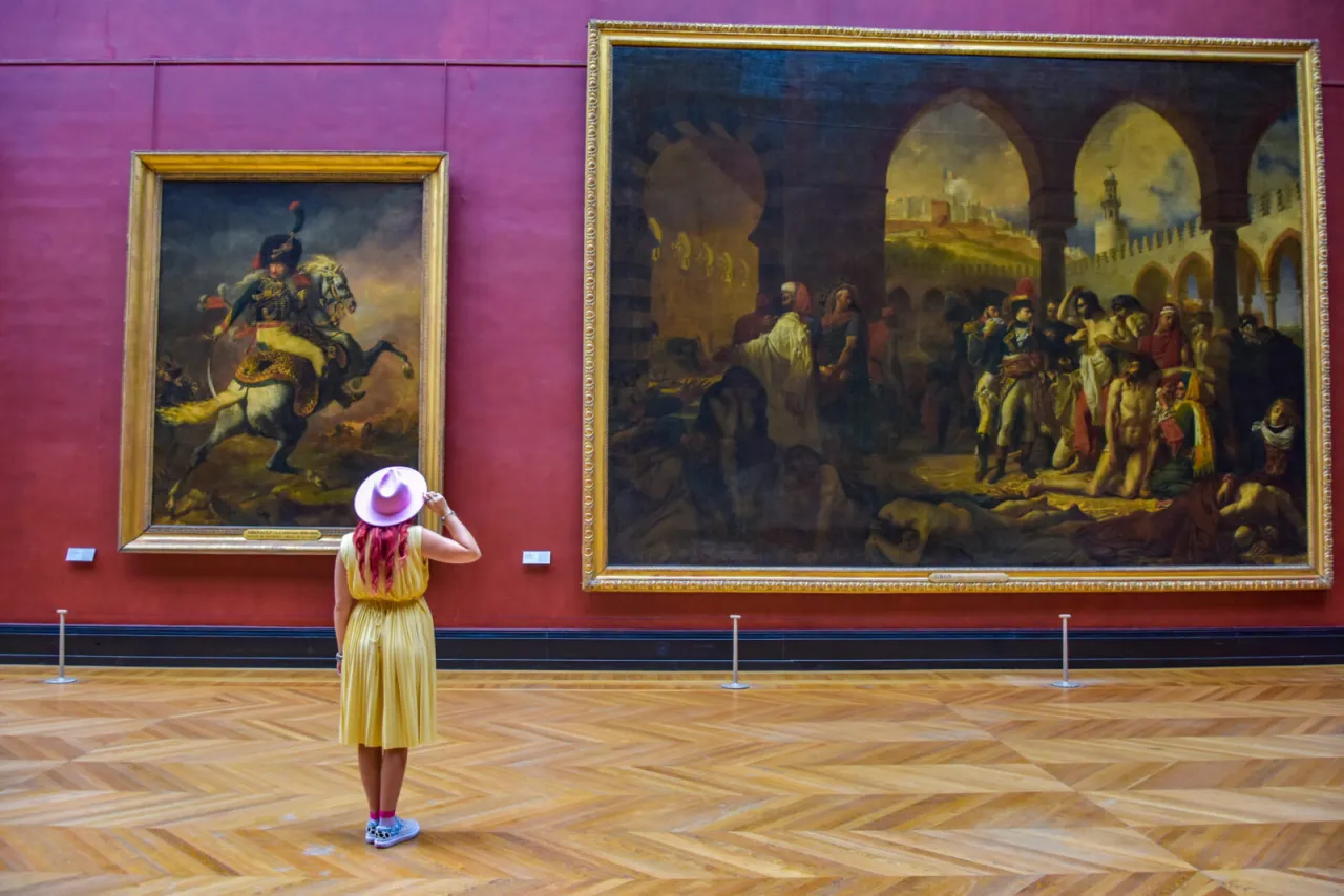 Kobieta w żółtej sukience i różowym kapeluszu podziwia obrazy w Luwrze. Idealny strój, by poczuć się jak na lekcji 