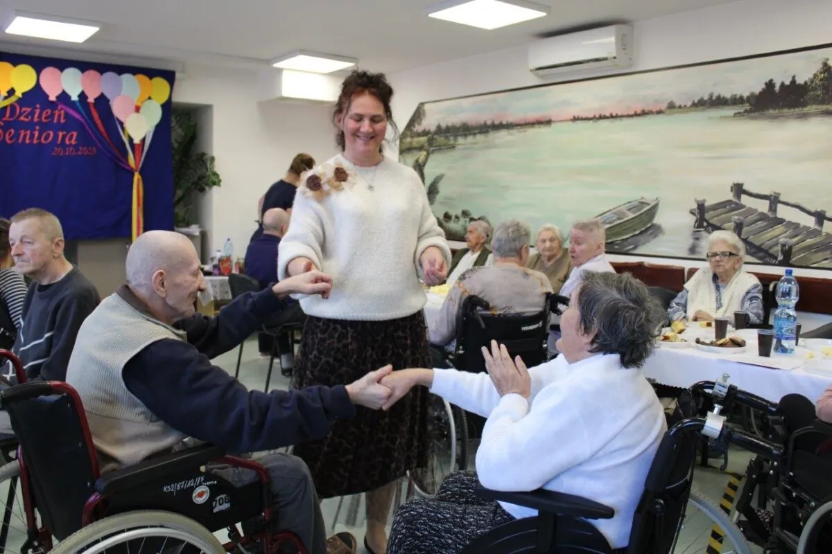 Radosny dzień seniora w domu opieki. Uśmiechnięta opiekunka tańczy z panem na w&oacute;zku, inni seniorzy obserwują.