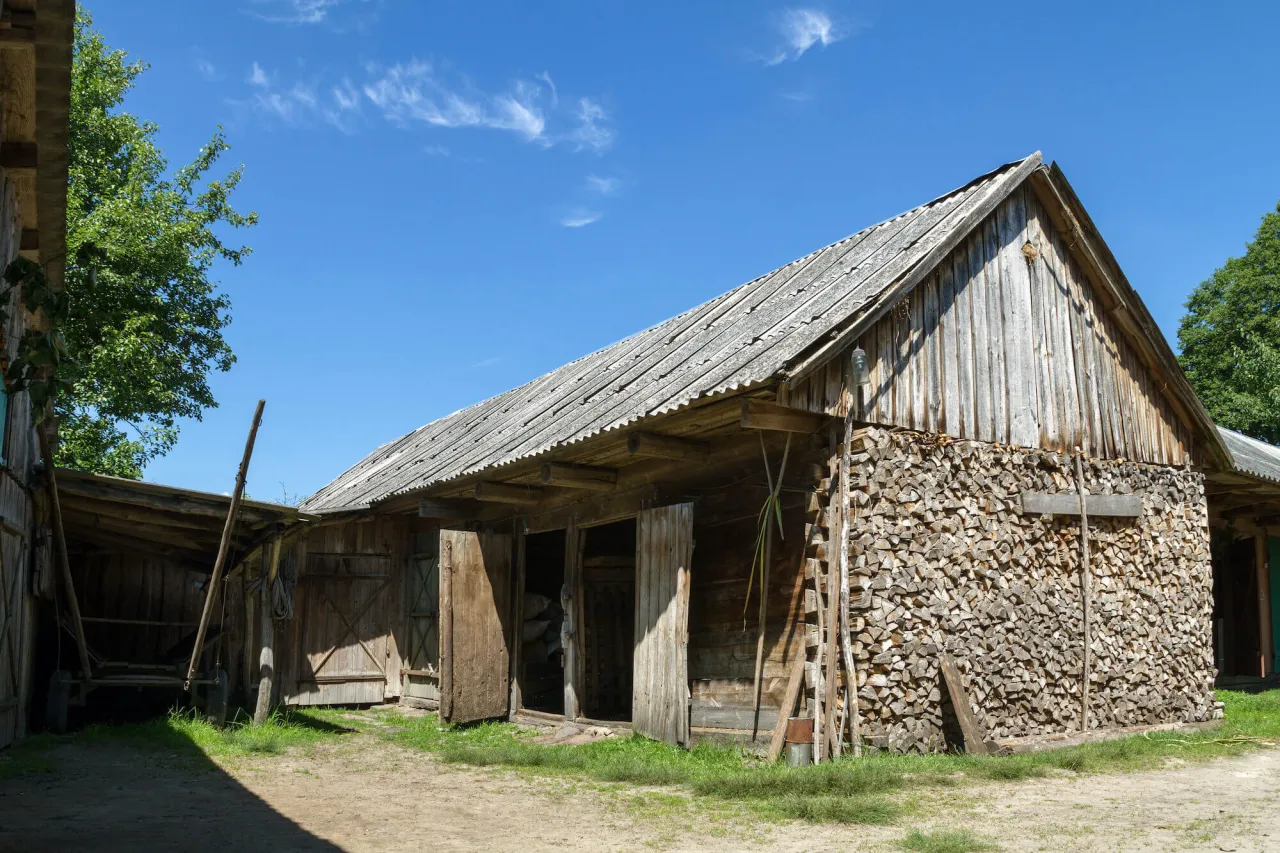 Drewniana stodoła, typowy budynek gospodarczy, z otwartymi drzwiami i stertą drewna opałowego.