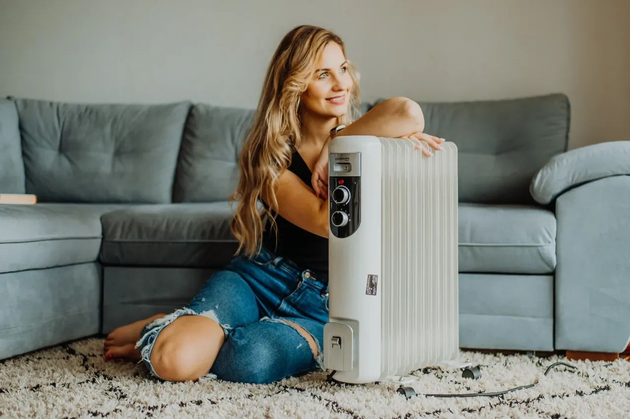 Uśmiechnięta kobieta siedzi na dywanie obok białego grzejnika olejowego, który zapewnia ciepło w chłodne dni.