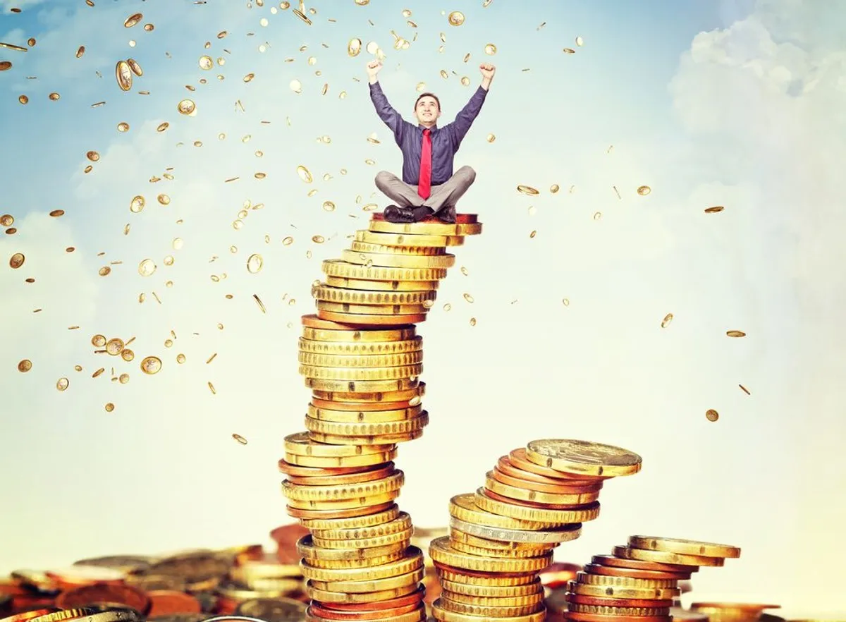 Mężczyzna siedzi na szczycie stosu monet, z rękami w górze. Czy dobra materialne czynią człowieka szczęśliwym?