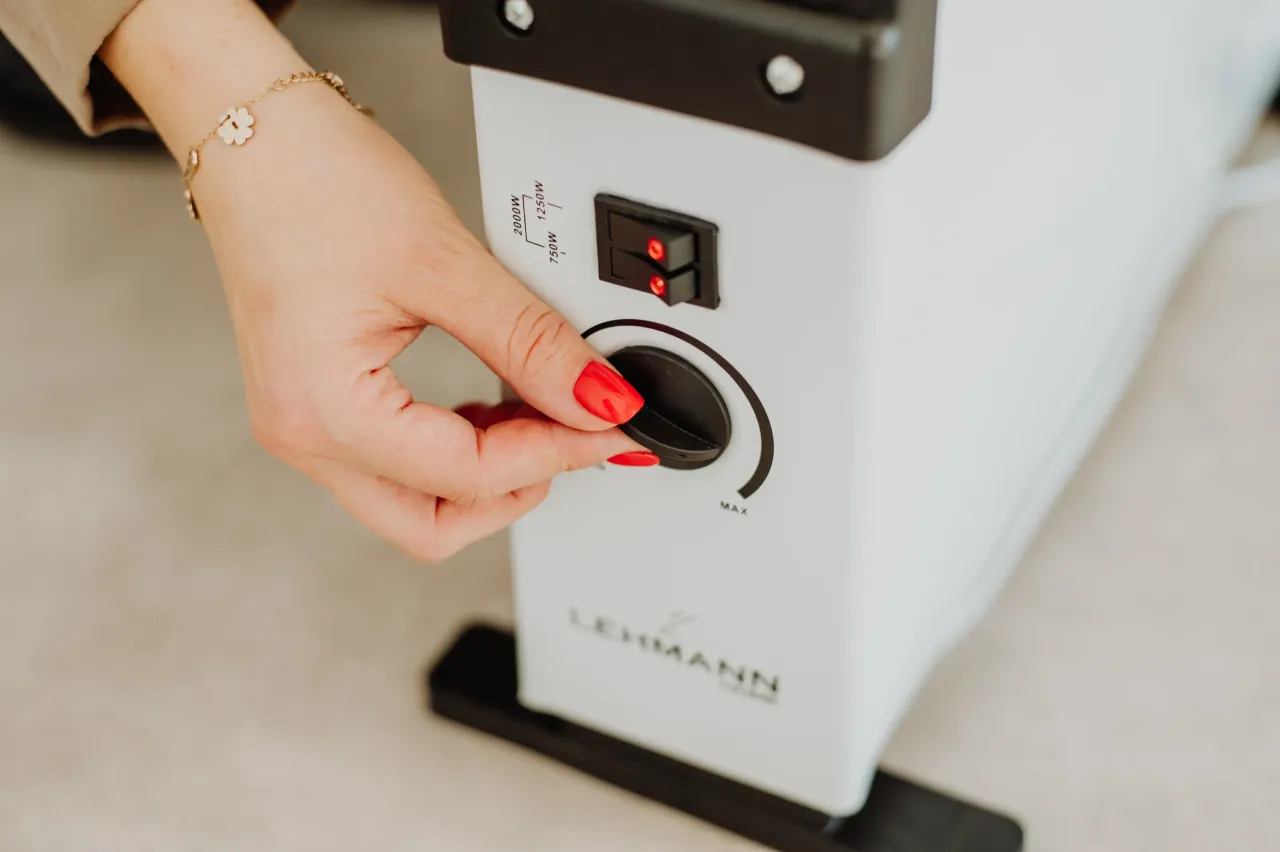 Dłoń z czerwoną hybrydą reguluje pokrętło grzejnika olejowego, wybierając moc grzania.