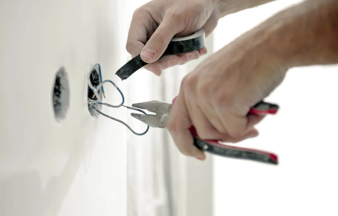 Elektryk naprawia instalację po przepięciu prądu. Używa taśmy izolacyjnej i kombinerki do przewod&oacute;w.