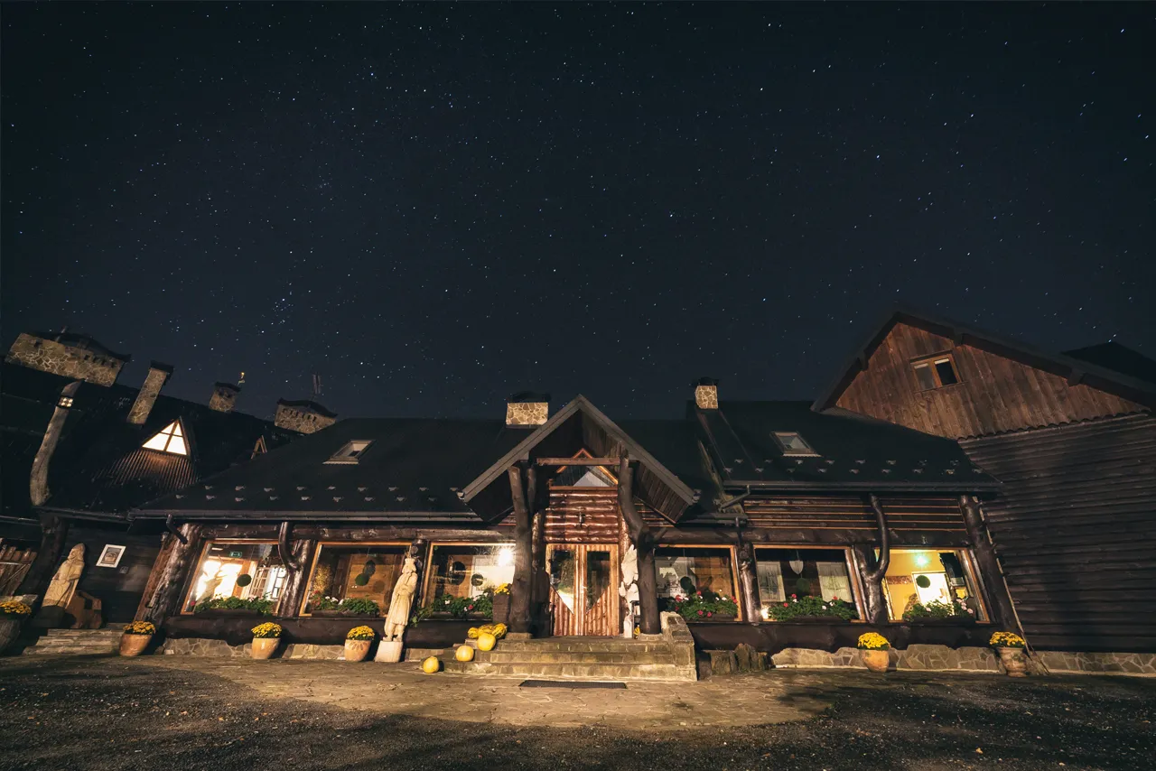 Nocne niebo pełne gwiazd nad drewnianą bac&oacute;wką Biały Jeleń. Ciepłe światło sączy się z okien, zapraszając do środka.