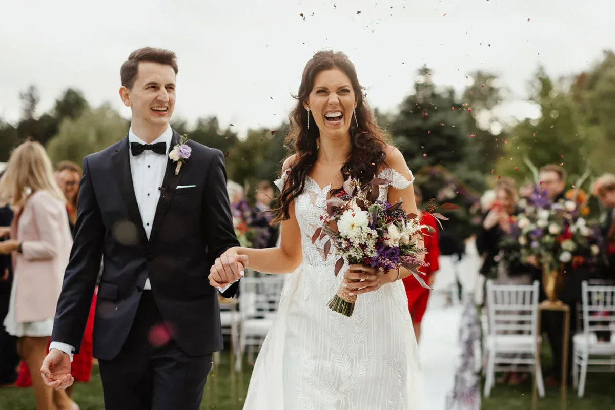 Szczęśliwa para młoda idzie alejką, obsypana płatkami. Idealne ujęcie, by pokazać, co napisać w mailu do fotografa ślubnego.