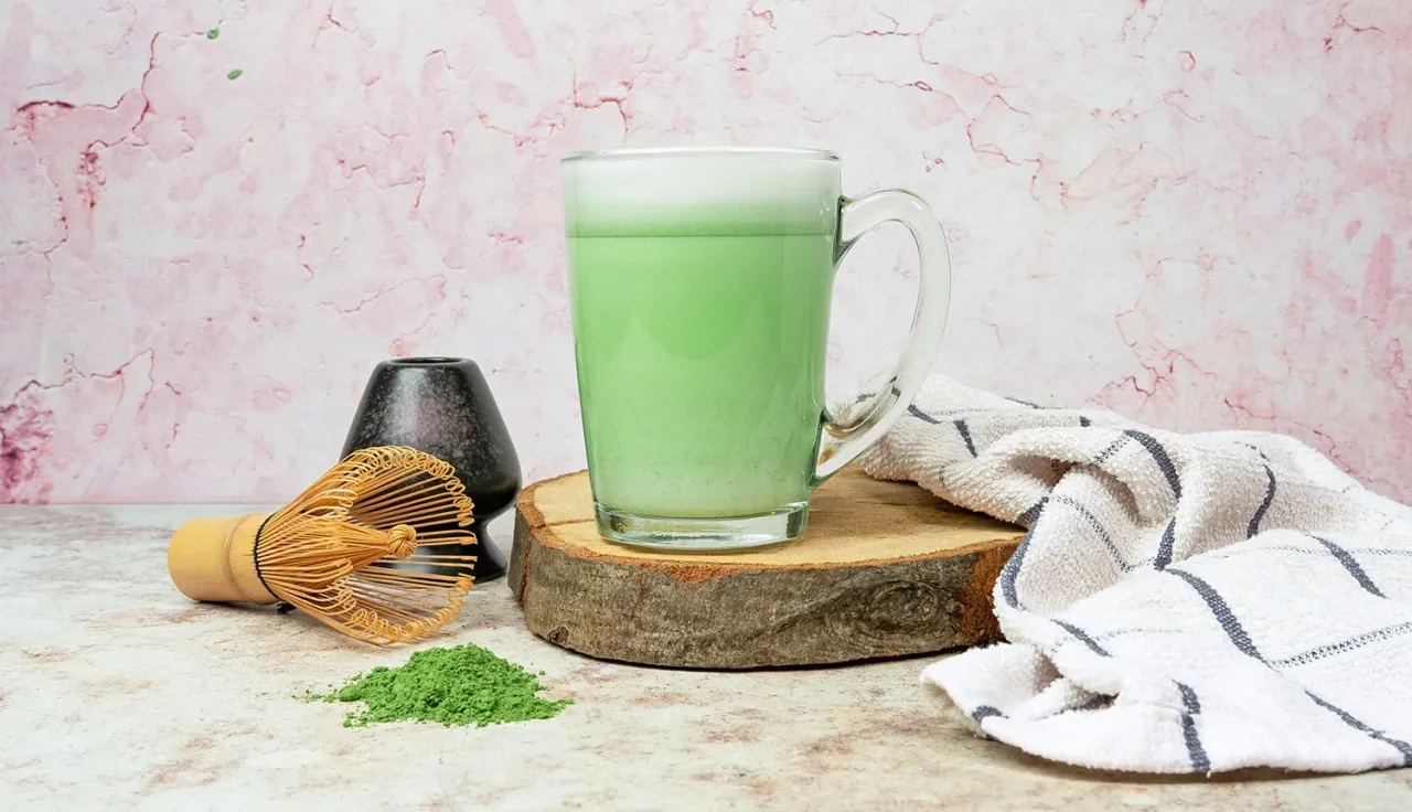 Zielony, spieniony matcha latte w szklance, obok bambusowa miotełka i proszek matcha. Idealny nap&oacute;j, jeśli zastanawiasz się, matcha latte gdzie kupić.