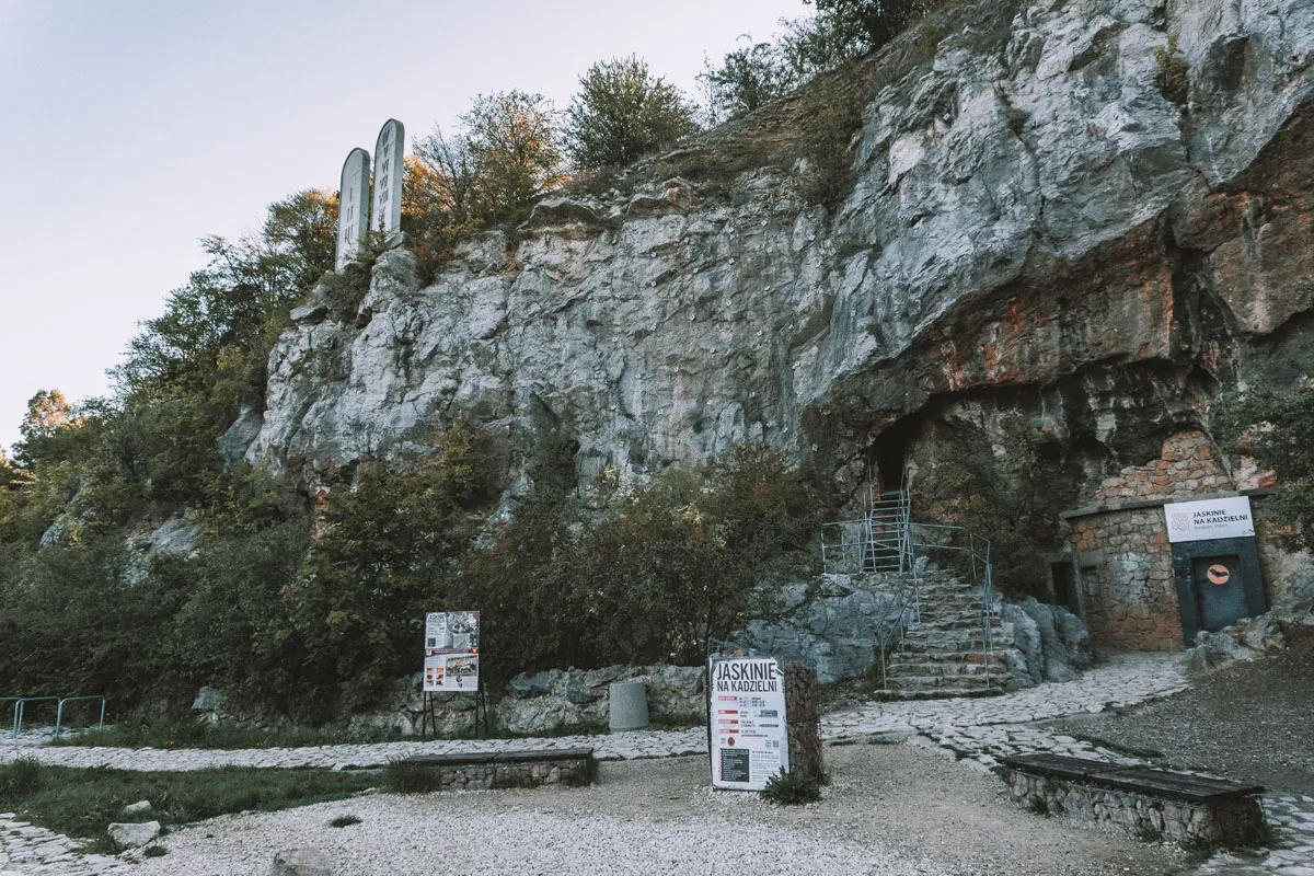 Kamieniste ścieżki prowadzą do wejścia do jaskiń w rezerwat przyrody Kadzielnia. Skalna ściana porośnięta zielenią, z tablicami informacyjnymi i ławkami.