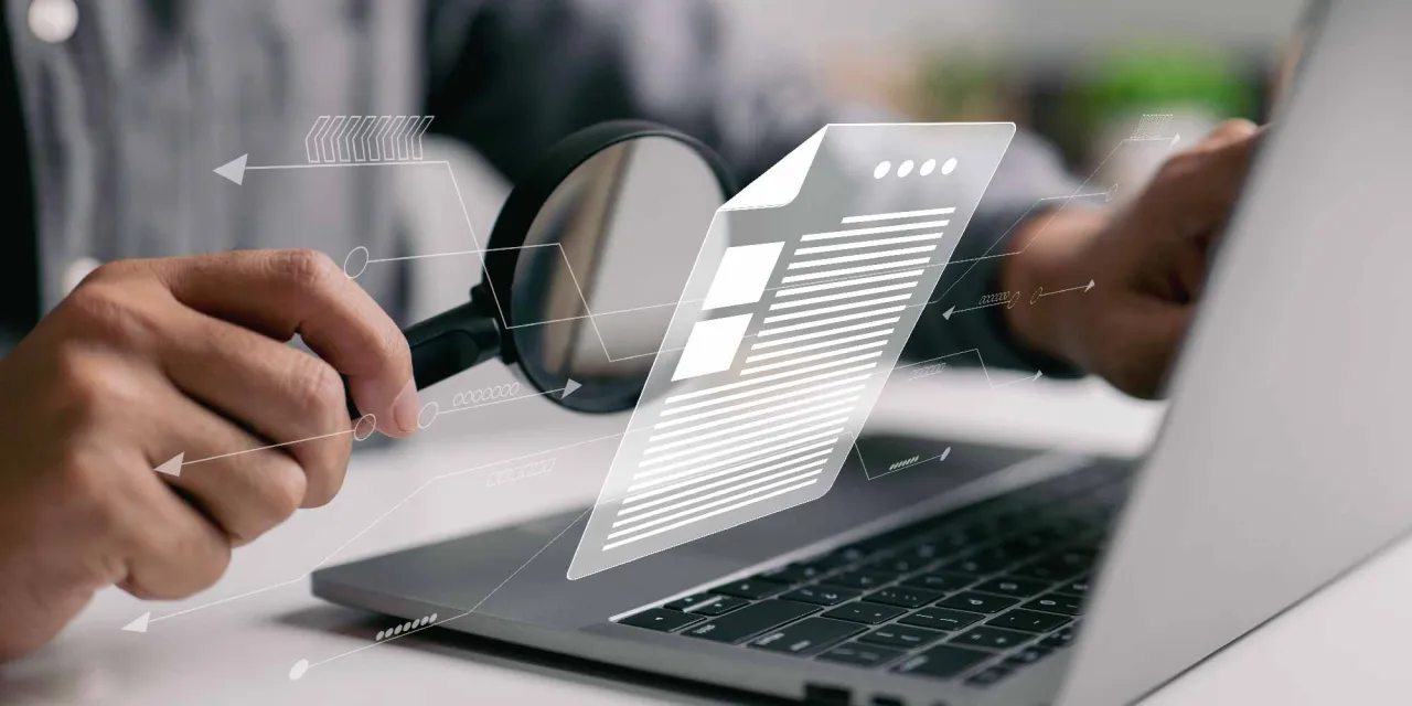 Ręka z lupą analizuje dokument na laptopie, szukając co to jest duplicate content.