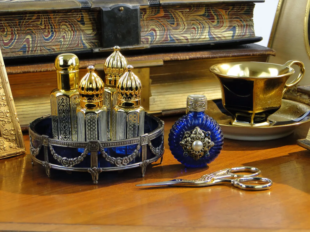 Złote i niebieskie flakoniki perfum, ozdobna filiżanka i stare księgi tworzą elegancką kompozycję.