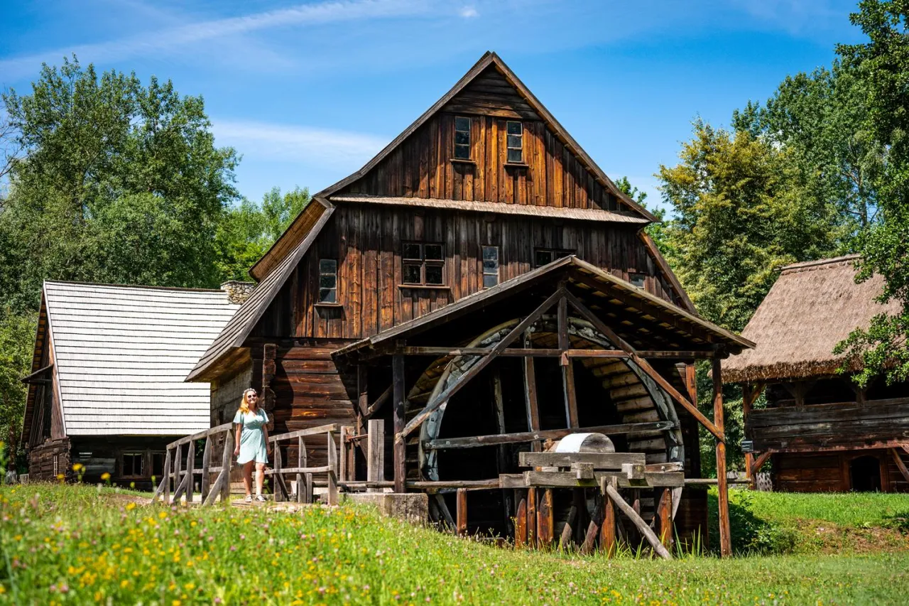 Drewniany młyn wodny i chata kryta strzechą to ciekawe miejsca, co można zwiedzić w Opolu. Kobieta w letniej sukience podziwia architekturę.