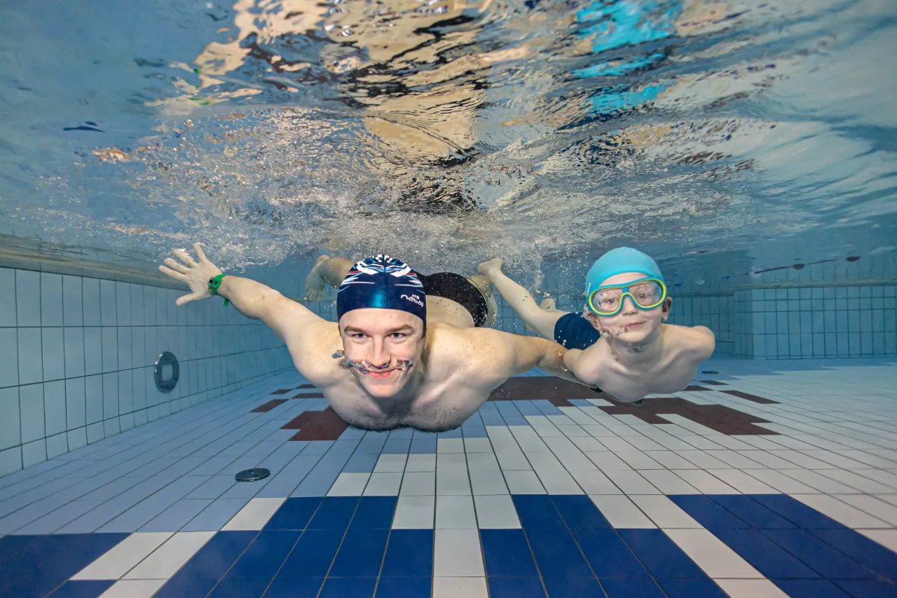 Dw&oacute;ch pływak&oacute;w, dorosły i dziecko, uśmiecha się pod wodą. To świetny przykład, ile trwa nauka pływania &ndash; radość i postępy widać od razu!