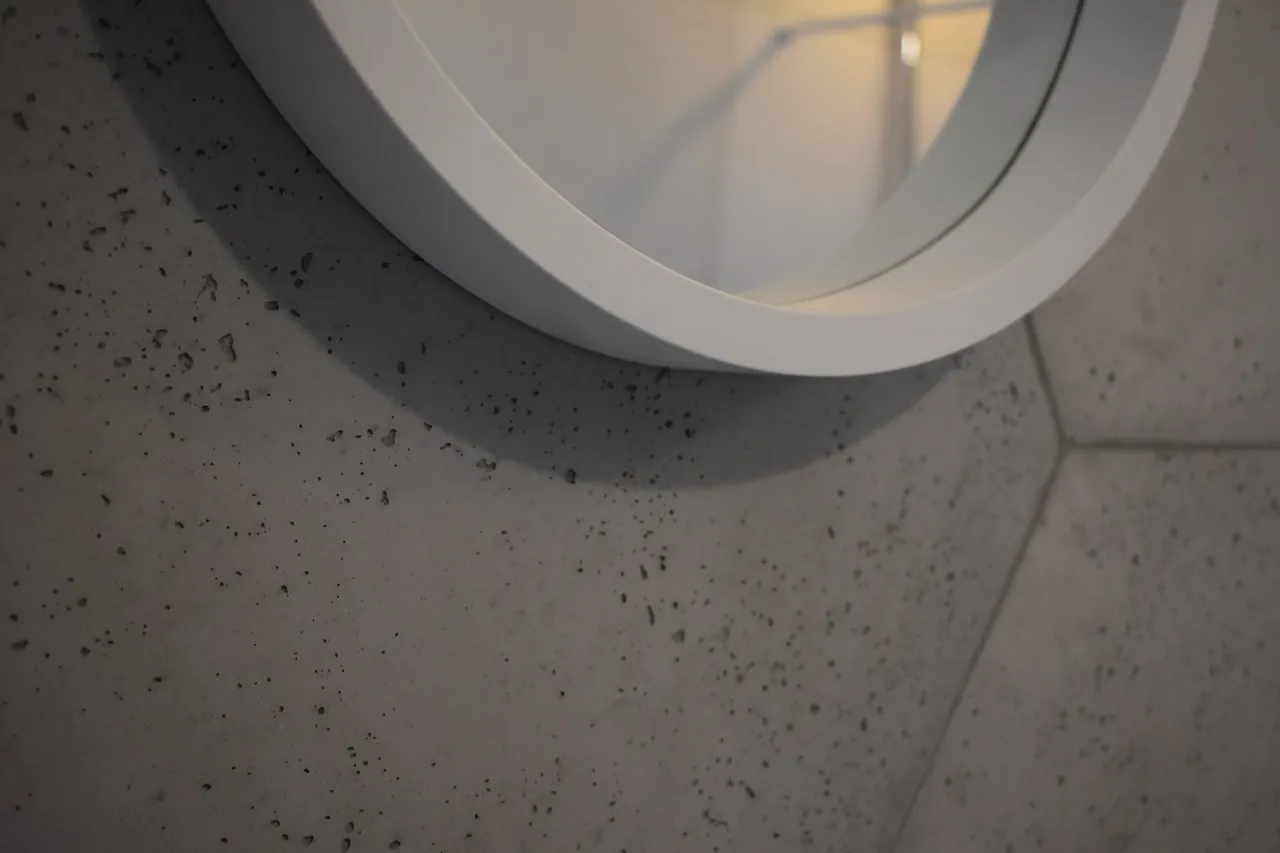 Nowoczesna ściana z betonu zbrojonego wł&oacute;knem szklanym, z widocznym fragmentem białej, okrągłej ramy lustra.