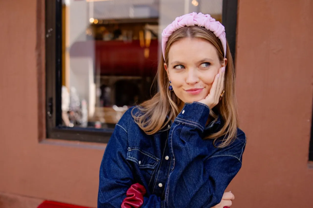 Uśmiechnięta, clean girl w dżinsowej kurtce i r&oacute;żowej opasce na głowie, opiera policzek na dłoni.