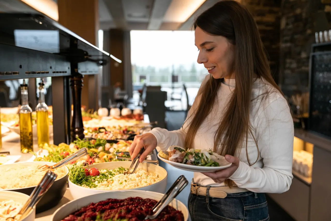 Uśmiechnięta kobieta nakłada sałatkę na talerz. Czy bon turystyczny można wykorzystać w restauracji? Tak, to świetny sposób na opłacenie posiłku!