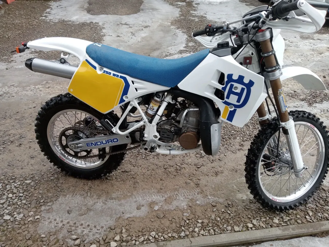 Klasyczny Husqvarna CR 250 Enduro gotowy na przygodę. Biało-żółta grafika i niebieskie siedzenie.