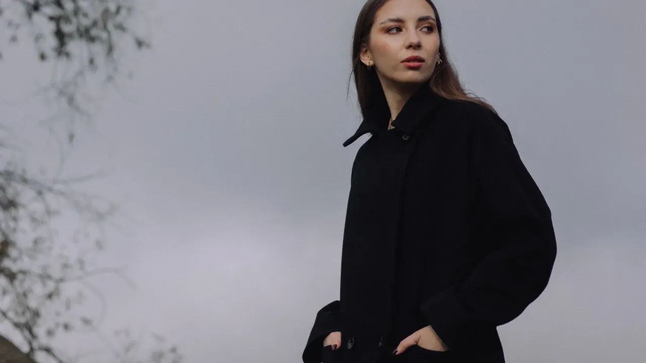 Młoda kobieta w czarnym płaszczu, z rękami w kieszeniach, spogląda w bok. Czy 18 stopni to ciepło?